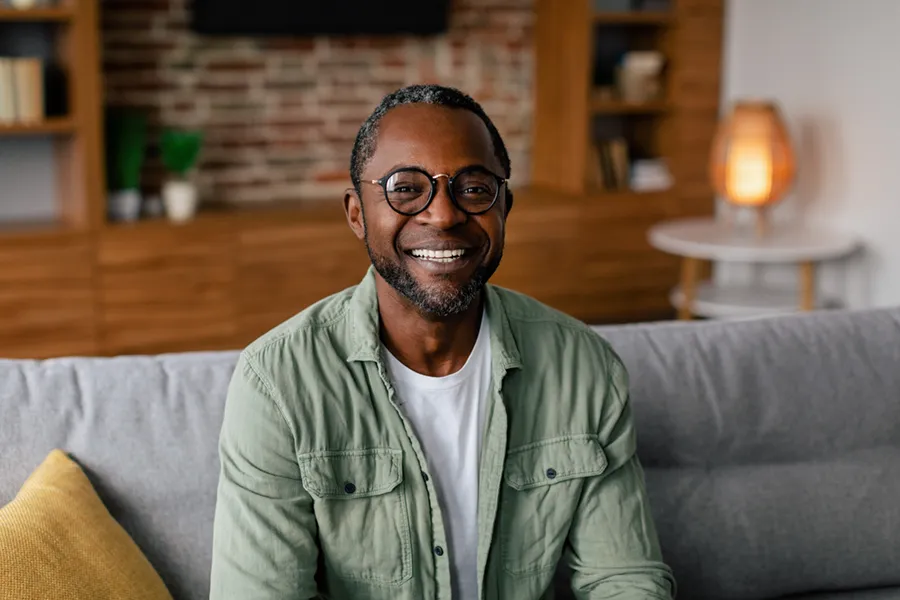 A middle aged black man in a green button up shirt sits on his couch smiling, happy with his treatment for andropause and low testosterone from Dr. Hadi Michael Rassael at Millennium Wellness in Bethesda-Chevy Chase.