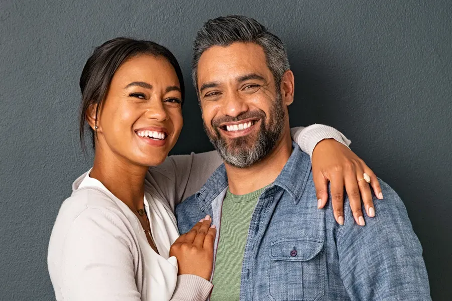 A middle-aged couple with their arms around each other standing against a gray background and smiling, happy with the bioidentical hormone therapy they received from Dr. Hadi Michael Rassael at Millennium Wellness in Bethesda-Chevy Chase.