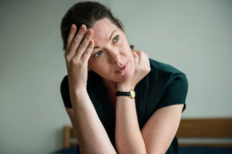 A woman in a dark blue dress sits on her bed holding her head in her hands with an exhausted expression. Get help for chronic fatigue syndrome from Dr. Hadi Michael Rassael at Millennium Wellness in [AERA].