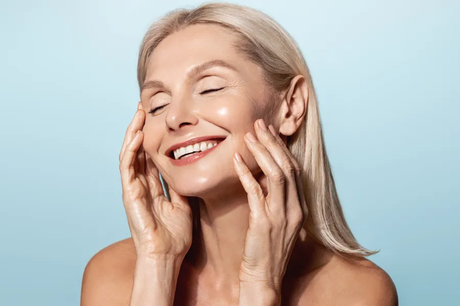 A mature blonde woman closes her eyes smiling and touches her smooth face. Get a facelift from Dr. Hadi Michael Rassael at Millennium Wellness in Bethesda-Chevy Chase.