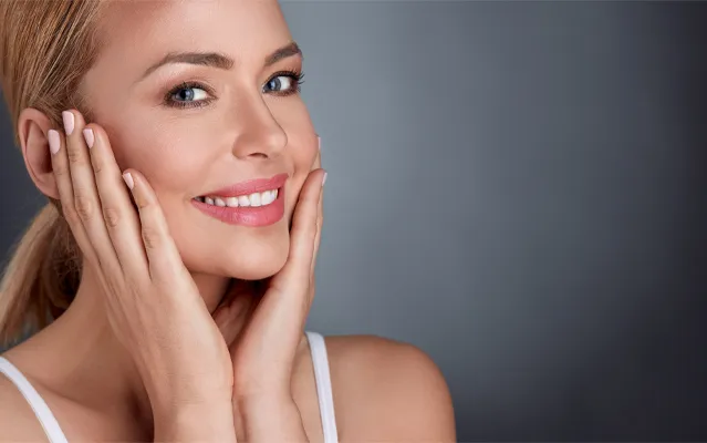 A middle-aged blonde woman with beautiful skin smiles and holds her face. Get facial treatments from Dr. Hadi Michael Rassael at Millennium Wellness in Bethesda-Chevy Chase.