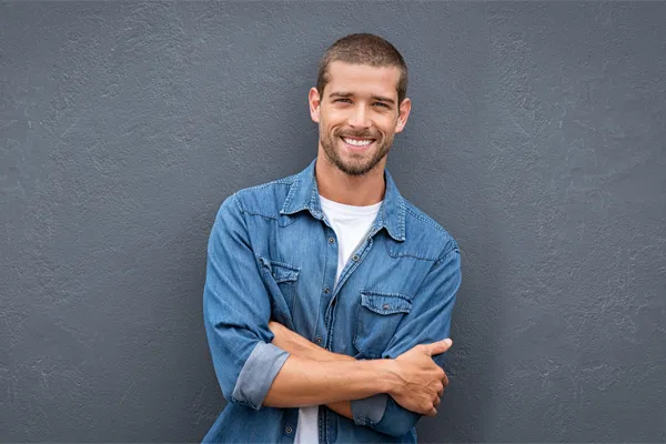 A man in a denim shirt stands smiling against a gray-blue wall, pleased with his testosterone hormone therapy from Dr. Hadi Michael Rassael at Millennium Wellness in Bethesda-Chevy Chase.