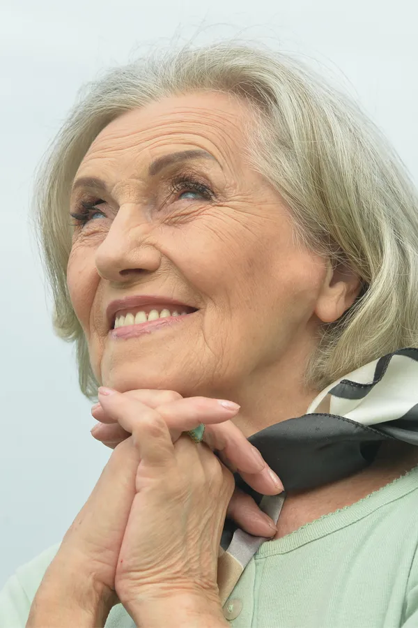 An older woman holds her hands together under her chin and looks up at a cool sky smiling. Get treatment for hot flashes and night sweats from Dr. Hadi Michael Rassael at Millennium Wellness in Bethesda-Chevy Chase.