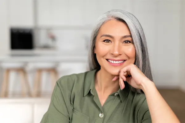 An asian woman with long gray hair and a green shirt sitting and smiling on her couch, happy with her menopause care from [NAME} in Bethesda-Chevy Chase.