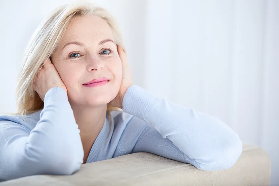 A mature blonde woman rests her head in her hands, smiling. Get a mini facelift from Dr. Hadi Michael Rassael at Millennium Wellness in Bethesda-Chevy Chase.