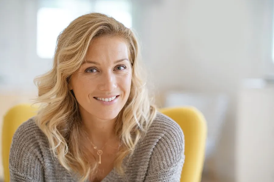 A blonde woman wearing a a gray sweater sits inside in a yellow chair, smiling and happy with her perimenopause treatment from Dr. Hadi Michael Rassael at Millennium Wellness in Bethesda-Chevy Chase.