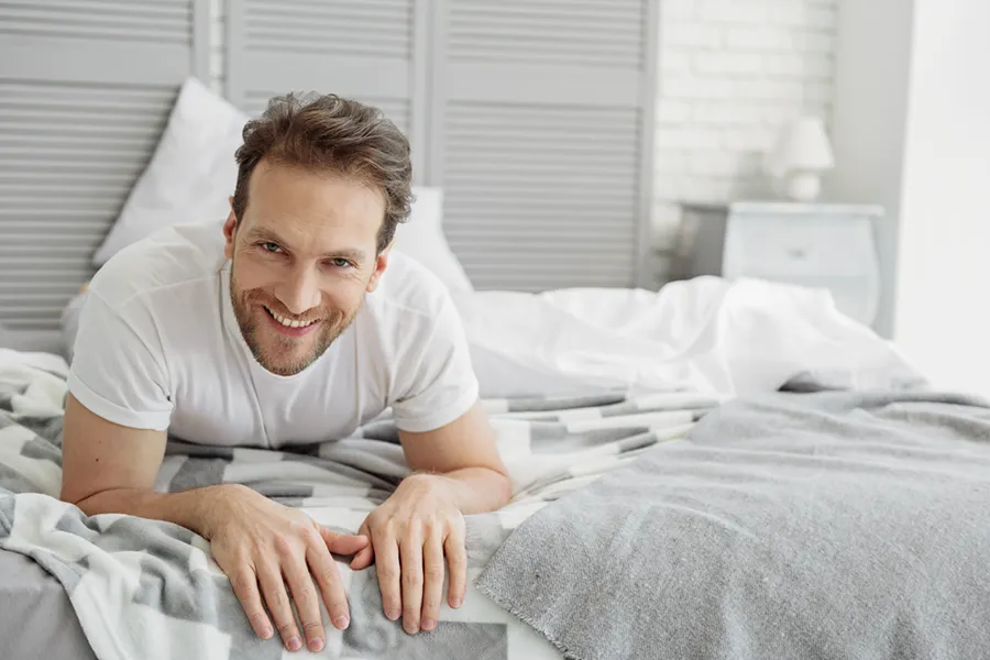 A man in a t-shirt lies on his stomach in bed smiling, happy with his sexual wellness treatments from Dr. Hadi Michael Rassael at Millennium Wellness in Bethesda-Chevy Chase.
