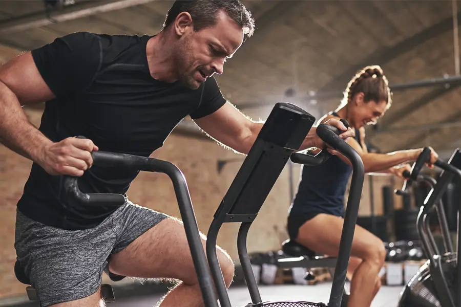 A couple in a brick gym work as hard as they can on exercise bikes, inspired by successful Tirzepatide treatment from Dr. Hadi Michael Rassael at Millennium Wellness in Bethesda-Chevy Chase.