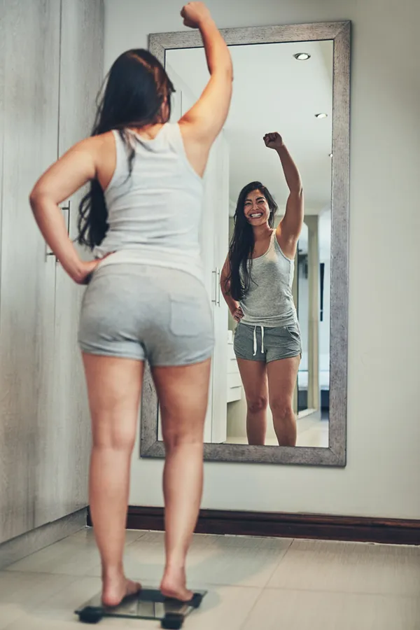 A woman in workout clothes stands on a scale looking in the mirror and pumping her hands in celebration of her successful Tirzepatide treatment from Dr. Hadi Michael Rassael at Millennium Wellness in Bethesda-Chevy Chase.
